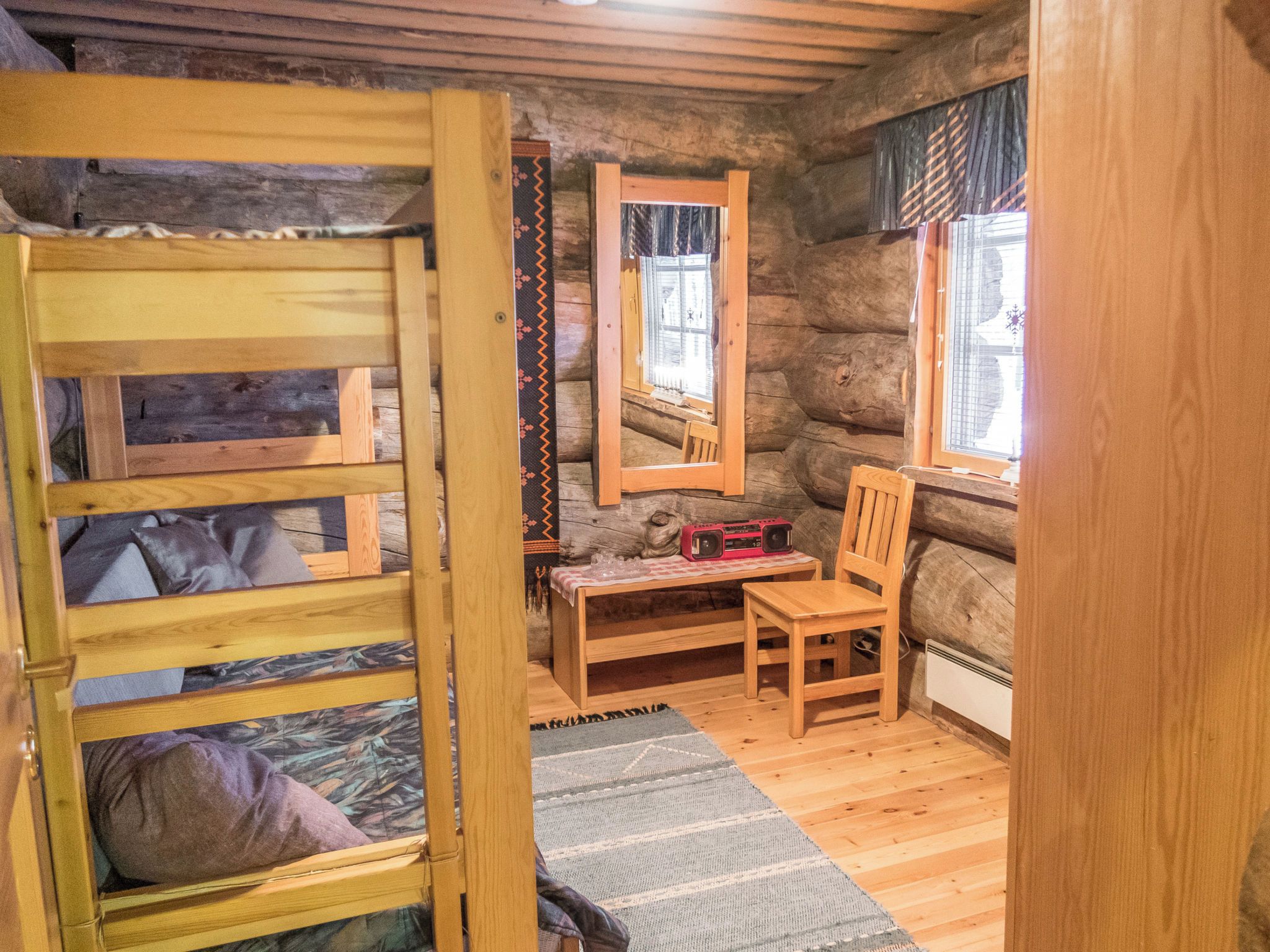 A rustic cabin interior with a bunk bed, a wooden table with a radio, a chair, and a window with curtains.