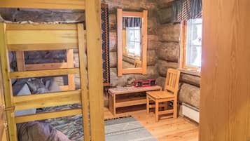 A rustic cabin interior with a bunk bed, a wooden table with a radio, a chair, and a window with curtains.