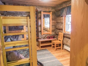 Interior rústico de cabaña con literas, una mesa de madera con una radio, una silla y una ventana con cortinas.
