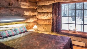 Une chambre à coucher aménagée dans un chalet en rondins, avec un lit, une fenêtre avec des rideaux et une vue sur une forêt enneigée.