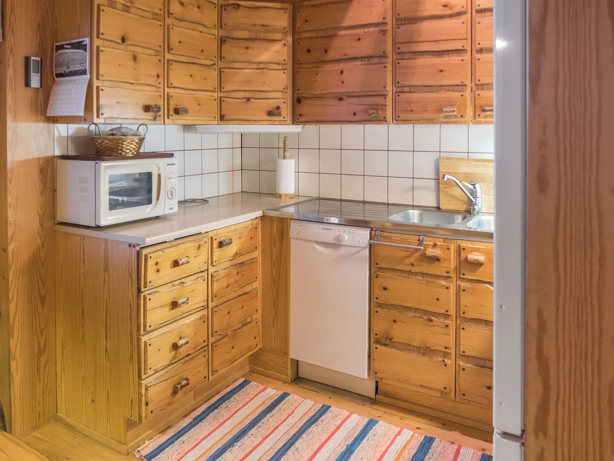 Ett kompakt kök med träskåp, en mikrovågsugn, en diskmaskin och en kakelbeklädd vägg bakom arbetsbänken.
