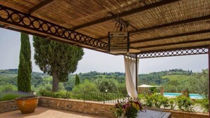 A covered patio with a wooden table and chairs, a large vase of flowers, and a view of a swimming pool and hills.