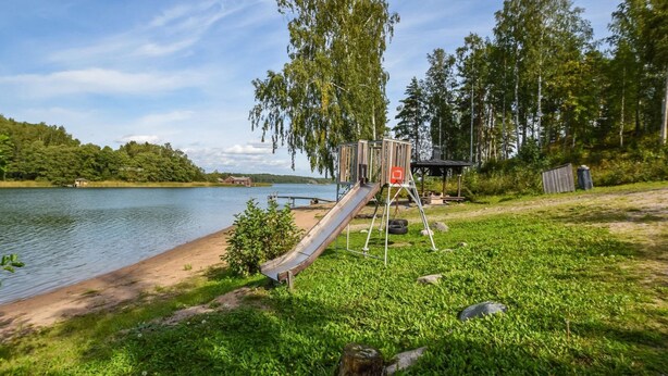 En legeplads med rutchebane og gyngestativ ved en sø, omgivet af træer og græs.