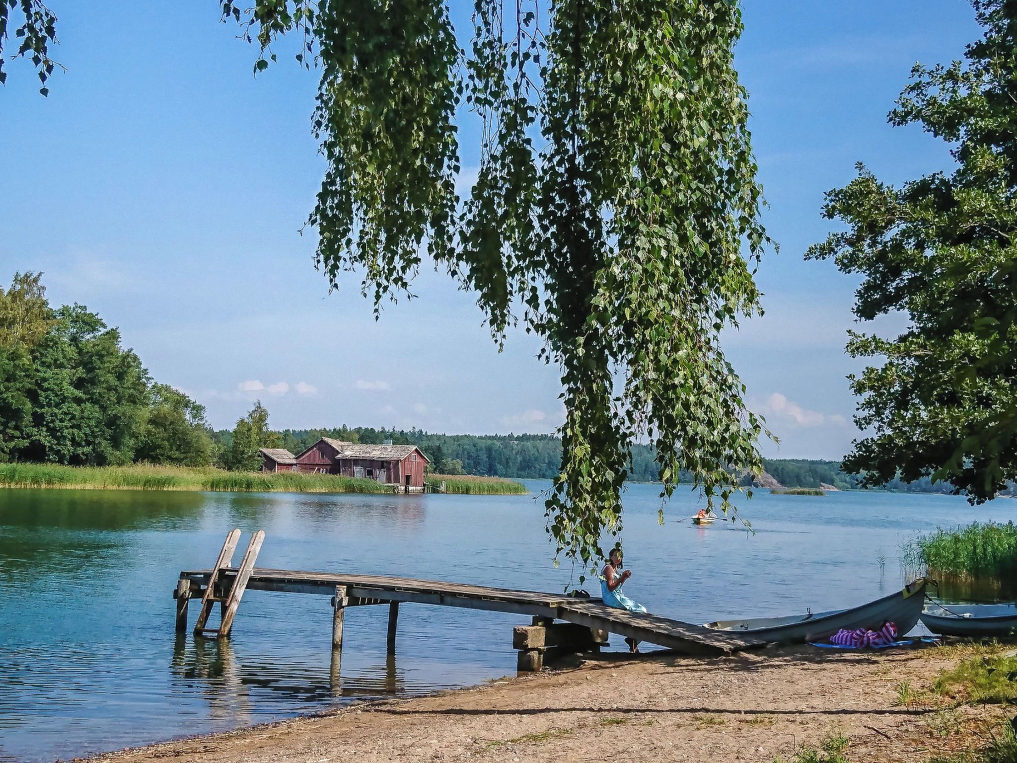 Une scène au bord d’un lac, avec une jetée en bois s’étendant dans l’eau, un bâtiment rouge en arrière-plan et un saule pleureur dont les branches pendent au-dessus de la jetée.
