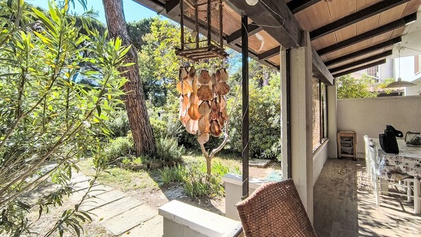 An outdoor patio with a wicker chair, a hanging decorative item, and a view of a garden with various plants and a tree. - Barracuda by Interhome (Grosseto)