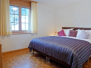 A bedroom with a wooden floor, a bed with a patterned bedspread, a window with curtains, and a bedside table with a lamp. - Chalet Perle by Interhome (Grindelwald)