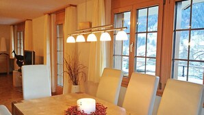 A dining area with a table set for four, large windows with a view of a snowy landscape, and a modern chandelier. - Chalet Perle by Interhome (Grindelwald)