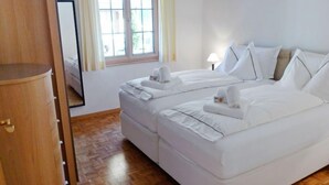 A hotel room with a wooden wardrobe, a double bed with white bedding, a window with curtains, and a wooden parquet floor. - Chalet Perle by Interhome (Grindelwald)