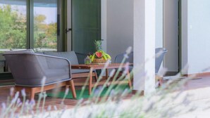Une chambre moderne avec une grande fenêtre, une table en bois sur laquelle se trouve un panier de fruits, et un canapé gris.