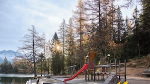 Un parc infantil amb un tobogan vermell i elements per escalar, envoltat d'arbres i un llac, amb muntanyes al fons.