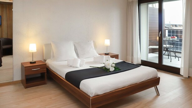 A modern hotel room with a large bed, two bedside tables with lamps, a view of the balcony through a sliding glass door, and a wooden floor. - Gamat 30 by Interhome (Villars)