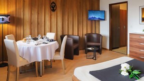 A hotel room with a wooden wall, a dining area with a round table and chairs, a bed with a black pillow, and a TV mounted on the wall. - Gamat 30 by Interhome (Villars)