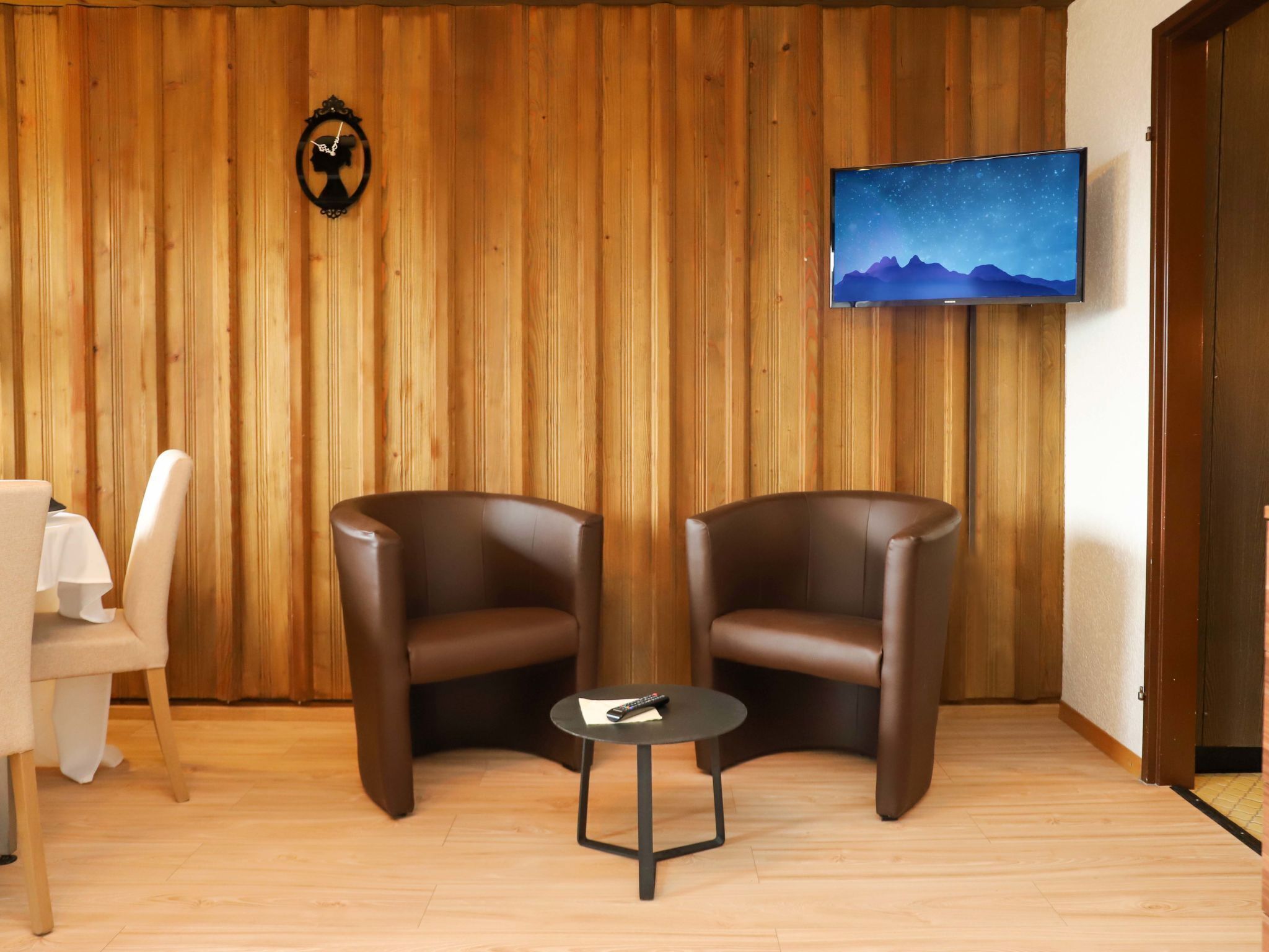 Ein Zimmer mit zwei Ledersesseln, einem kleinen runden Tisch, einem weißen Tisch mit weißer Tischdecke, einem an der Wand montierten Fernseher, der eine Berglandschaft zeigt, und einer Wand aus Holzpaneelen.