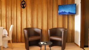 A room with two leather armchairs, a small round table, a white table with a white tablecloth, a wall-mounted TV displaying a mountain scene, and a wooden panel wall. - Gamat 30 by Interhome (Villars)