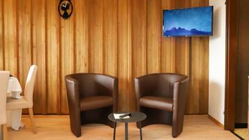 Ein Zimmer mit zwei Ledersesseln, einem kleinen runden Tisch, einem weißen Tisch mit weißer Tischdecke, einem an der Wand montierten Fernseher, der eine Berglandschaft zeigt, und einer Wand aus Holzpaneelen.