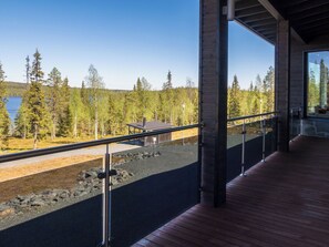 Un edificio moderno con una terraza de madera y barandilla de vidrio que da a un bosque y un lago.