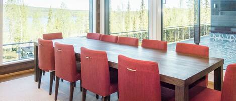 A modern dining room with a large wooden table and red upholstered chairs, large windows offering a view of trees, and a patterned floor.