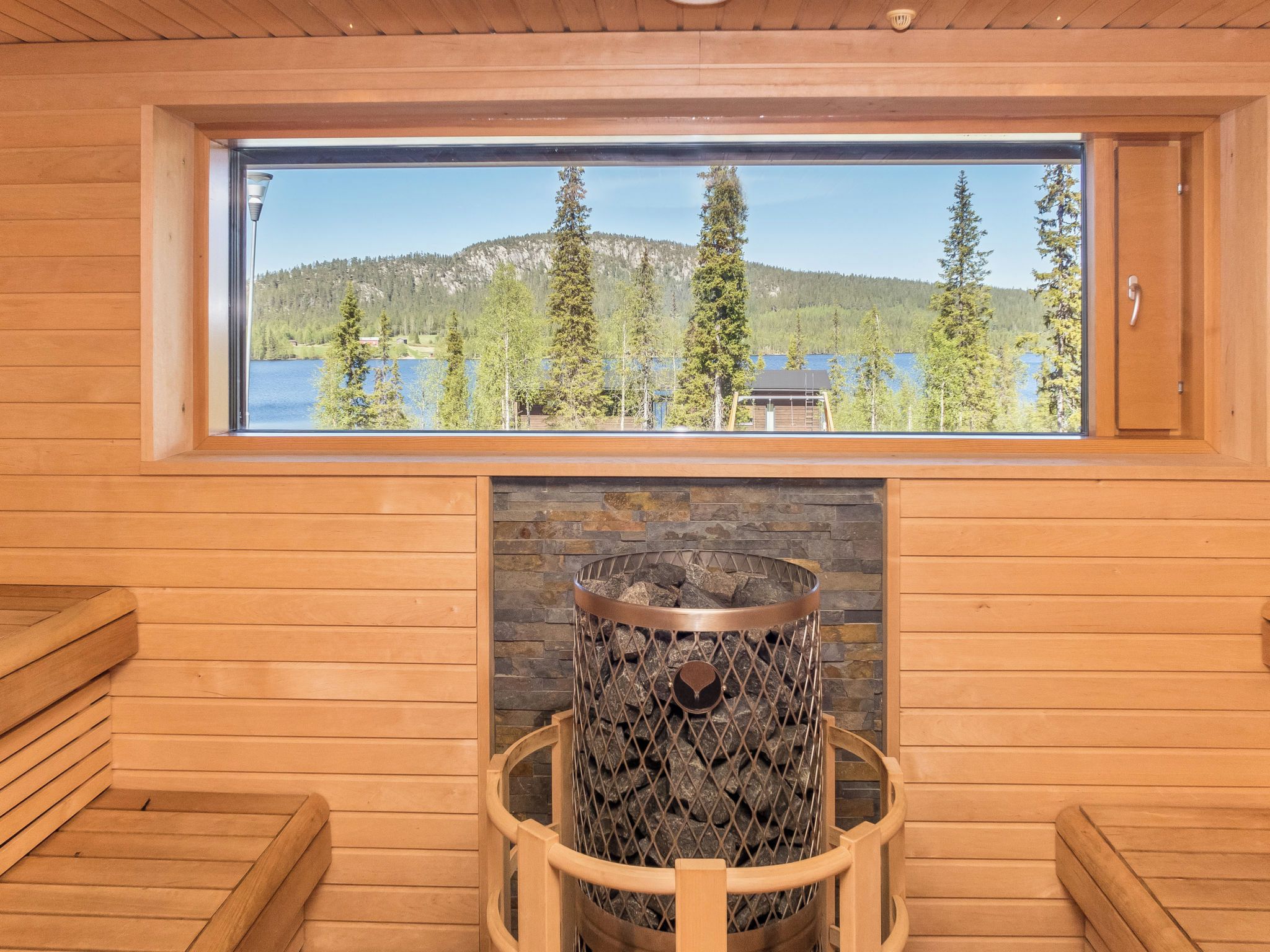 A wooden cabin interior with a stone fireplace, a window overlooking a lake and forest, and wooden benches.