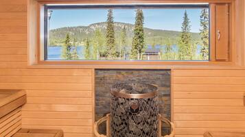 A wooden cabin interior with a stone fireplace, a window overlooking a lake and forest, and wooden benches.