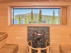 L’intérieur d’un chalet en bois est doté d’une cheminée en pierre, d’une fenêtre donnant sur un lac et une forêt, ainsi que de bancs en bois.