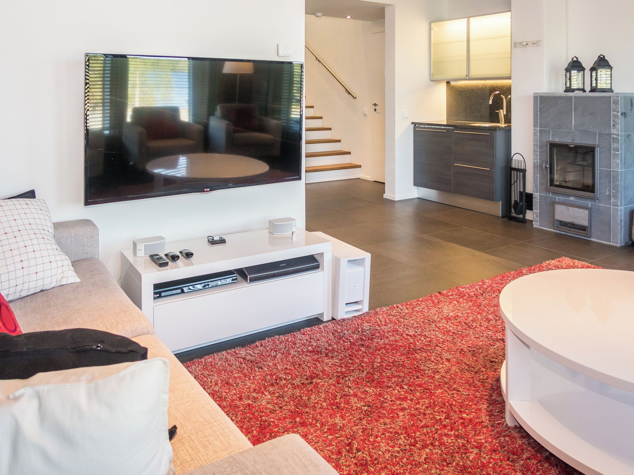 A modern living room with a flat-screen TV, a white entertainment unit, a red carpet, and a kitchen area with a fireplace.
