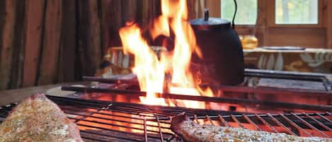 A grill with two pieces of meat cooking over an open flame, with a pot on a separate burner in the background.