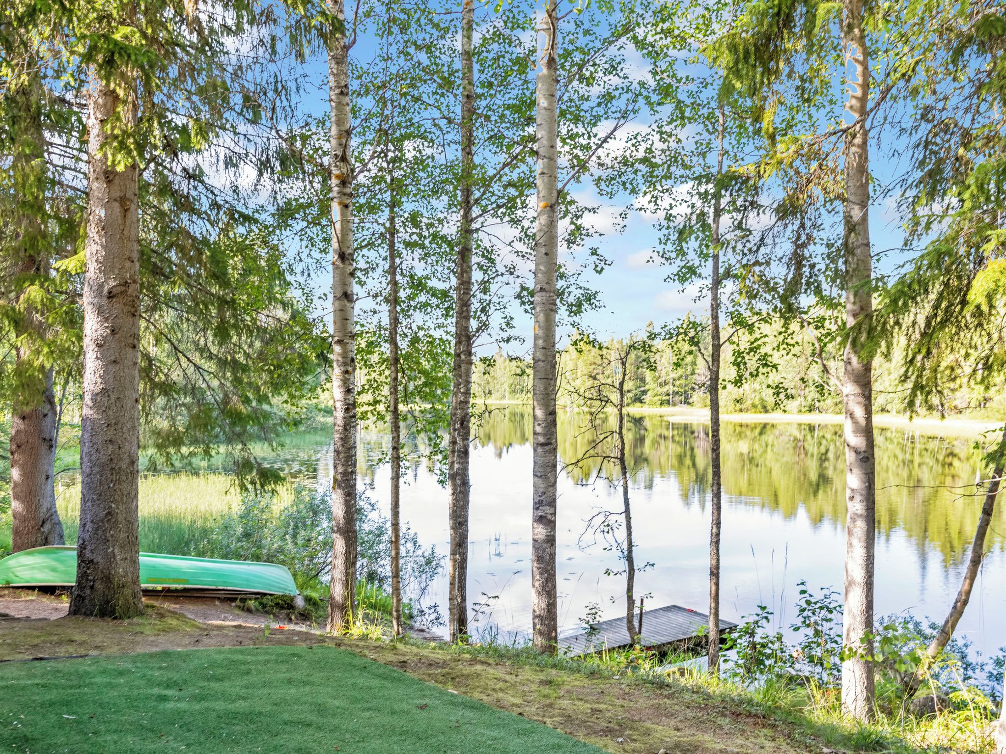 Un entorno tranquilo junto al lago con un bote verde amarrado cerca de la orilla, rodeado de altos árboles y una zona verde bien cuidada.