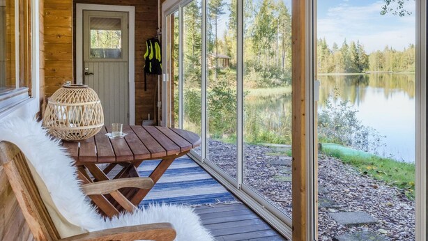 Une cabane en bois dotée d’une grande fenêtre donnant sur un lac, d’une table en bois et d’un banc recouvert d’un plaid en fourrure.