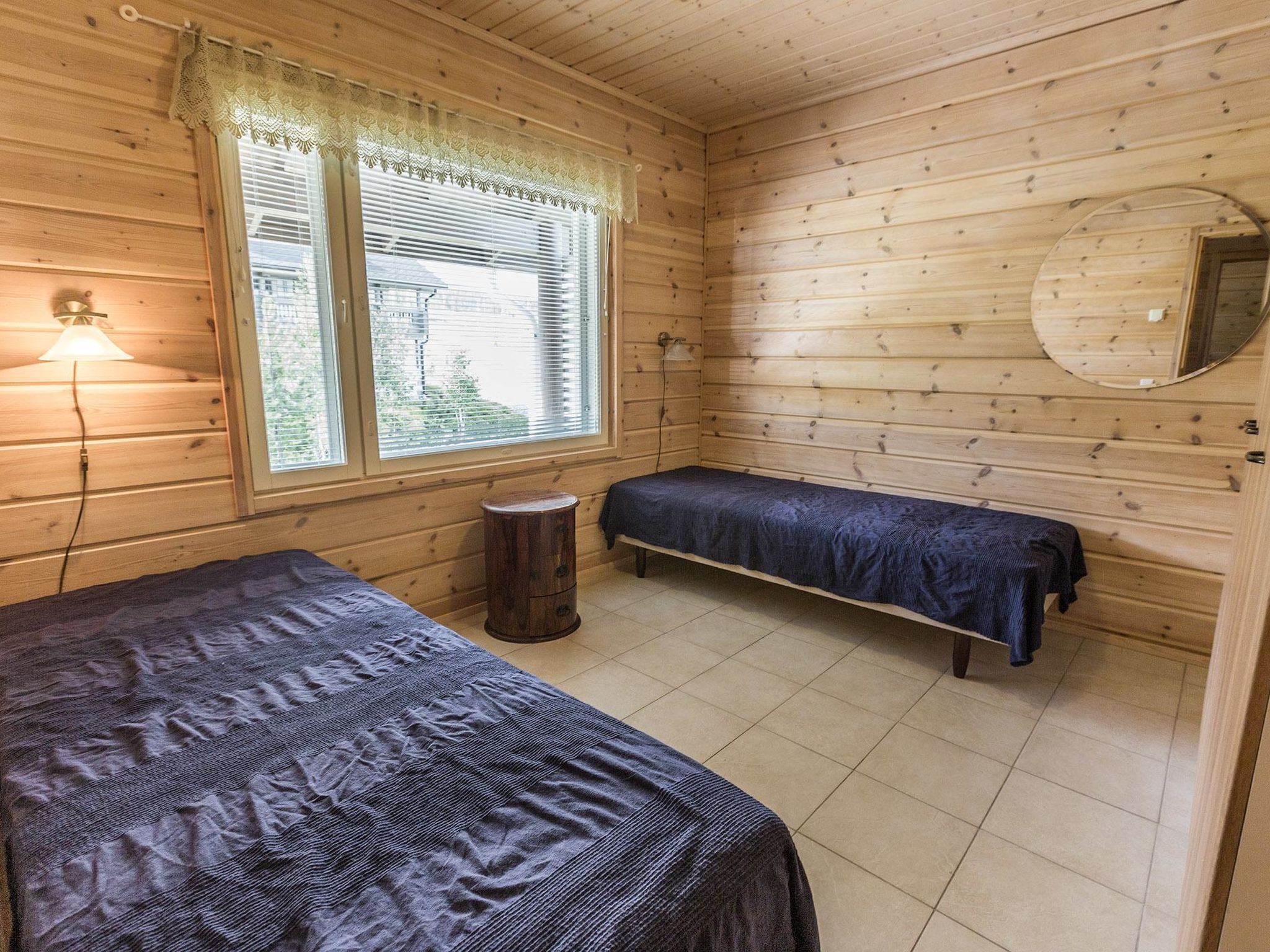 Una cabaña con dos camas, un taburete pequeño, una ventana con persianas, una lámpara y un espejo en la pared.