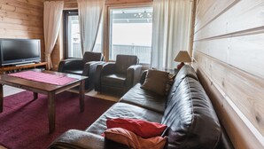 Ein gemütliches Blockhaus-Interieur mit Holzvertäfelung, einer Couch, zwei Sesseln, einem Fernseher und Balkonblick.