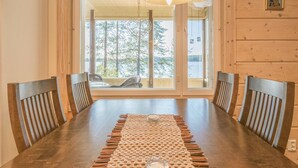 A dining area with a wooden table, four chairs, a woven placemat, and a view of trees through large windows. - Kärkiniemi by Interhome (Tuusniemi)
