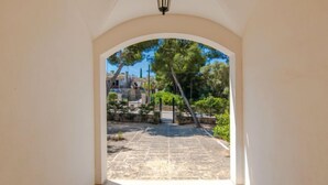 Un arco de entrada da a un patio con suelo de piedra, rodeado de vegetación y edificios.