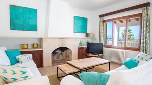 Un salón con chimenea, sofás blancos, cojines color turquesa, un televisor y vistas al exterior.