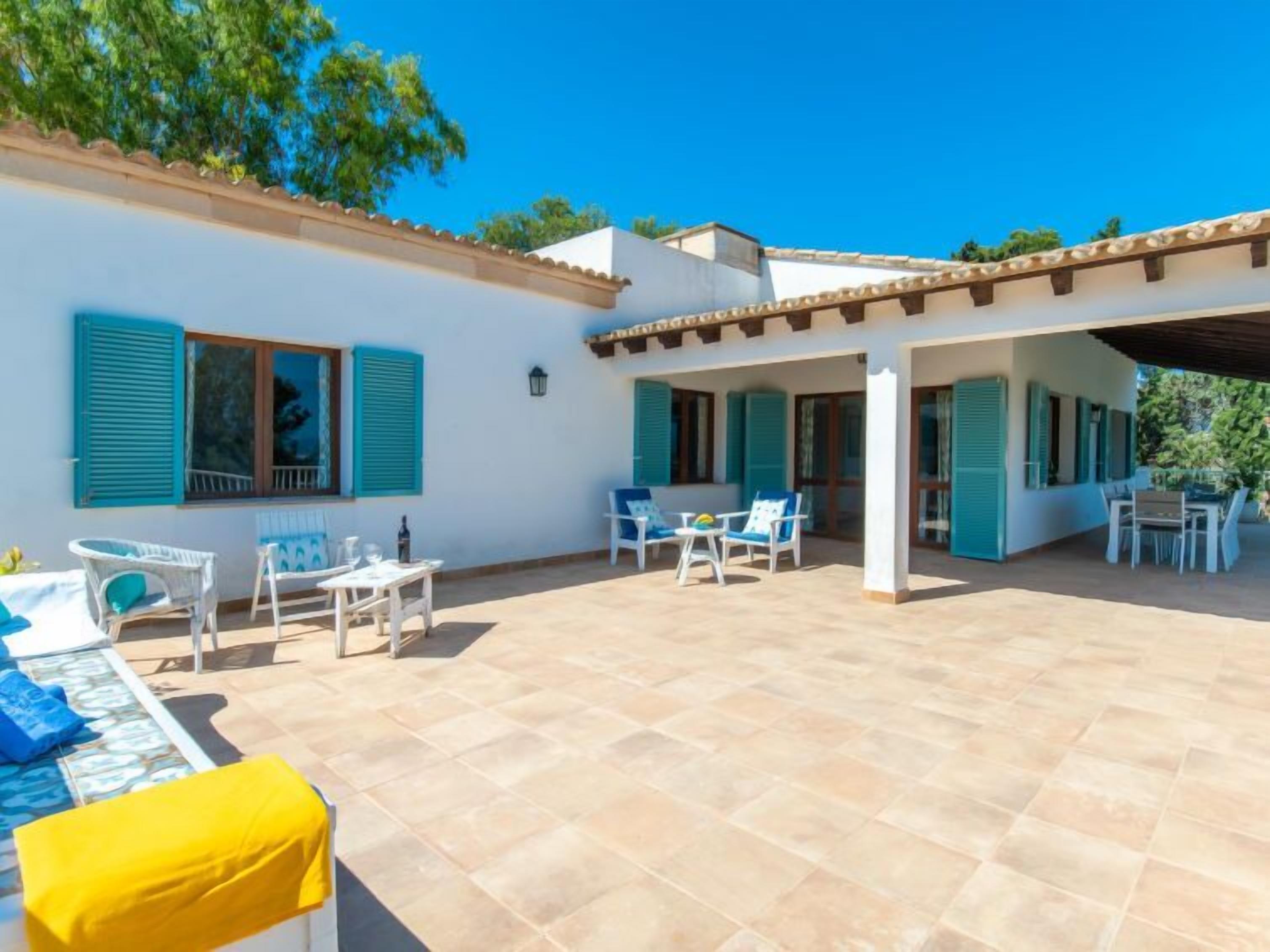 A covered outdoor patio with tiled flooring, white furniture, and blue shutters on the windows.