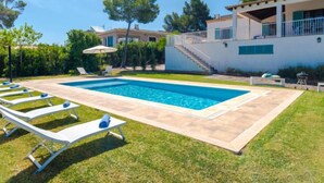 Piscina rectangular con sillones de descanso y una mesa, rodeada por un césped cuidado y un edificio de dos pisos con balcón.