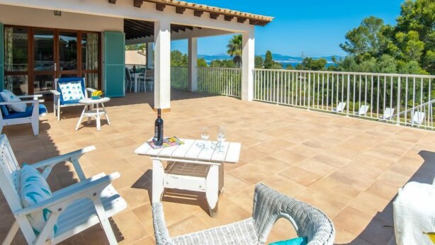 Un patio exterior cubierto con mobiliario de mimbre blanco, una mesa con una botella y vasos, y vistas al mar y a los árboles.