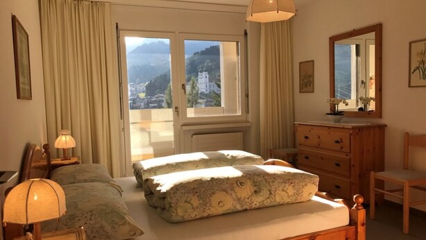 A bedroom with a bed, a dresser, a chair, a lamp, a mirror, and a window with a view of a mountain and buildings. - Disentiserhof Hönig by Interhome (Disentis)