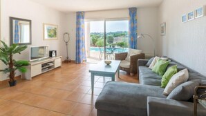 Un soggiorno spazioso con un ampio divano grigio, una televisione e un tavolo da pranzo. Si apre su una piscina e del verde attraverso le finestre.
