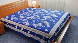 Een slaapkamer met een houten hoofdbord, een blauw beddegoed met witte patronen, een bedlampje en een raam met zonwering.