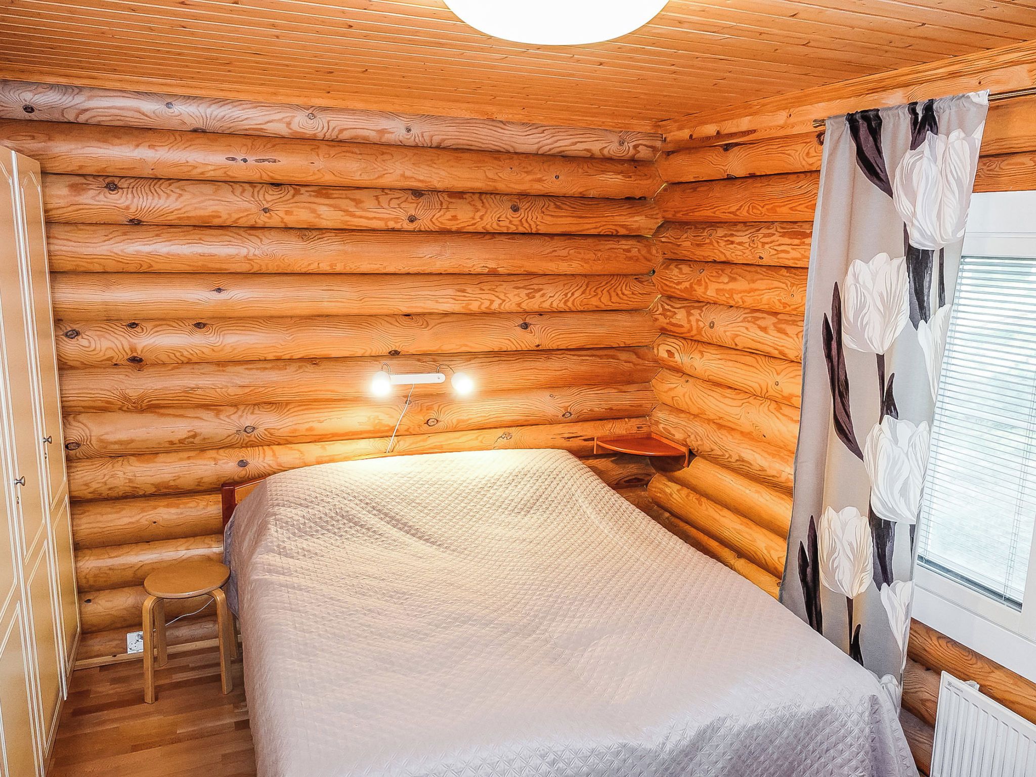 A log cabin room with a bed, a small stool, and a window with curtains.