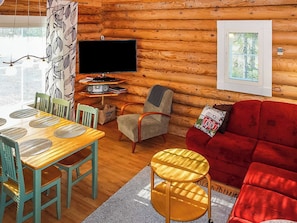 Een knus houten interieur met een wand van boomstammen, een eettafel met stoelen, een rode bank, een televisie en een raam met zonwering.