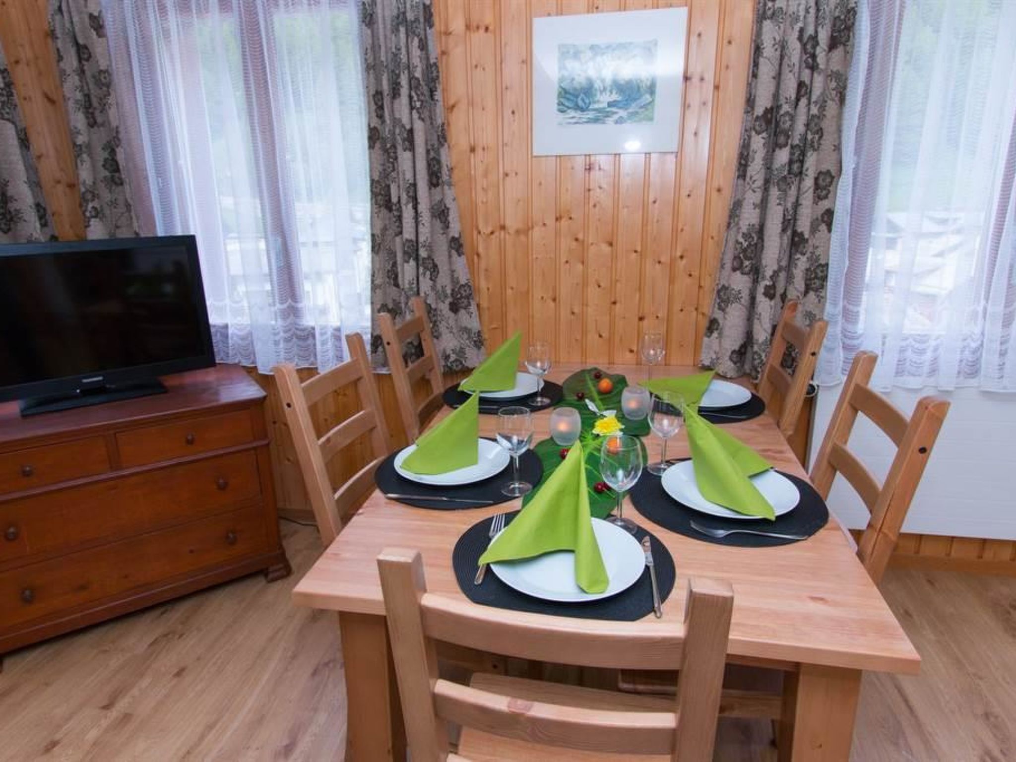 A dining area with a wooden table set for four, surrounded by chairs. A television is placed on a wooden cabinet to the left.