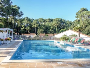 Ein rechteckiger Swimmingpool mit Fliesen und einer sonnenbeschienenen Liegefläche, umgeben von einem weißen Zaun. Auf der linken Seite befinden sich Liegestühle und eine kleine Struktur mit Pflanzen in Töpfen.