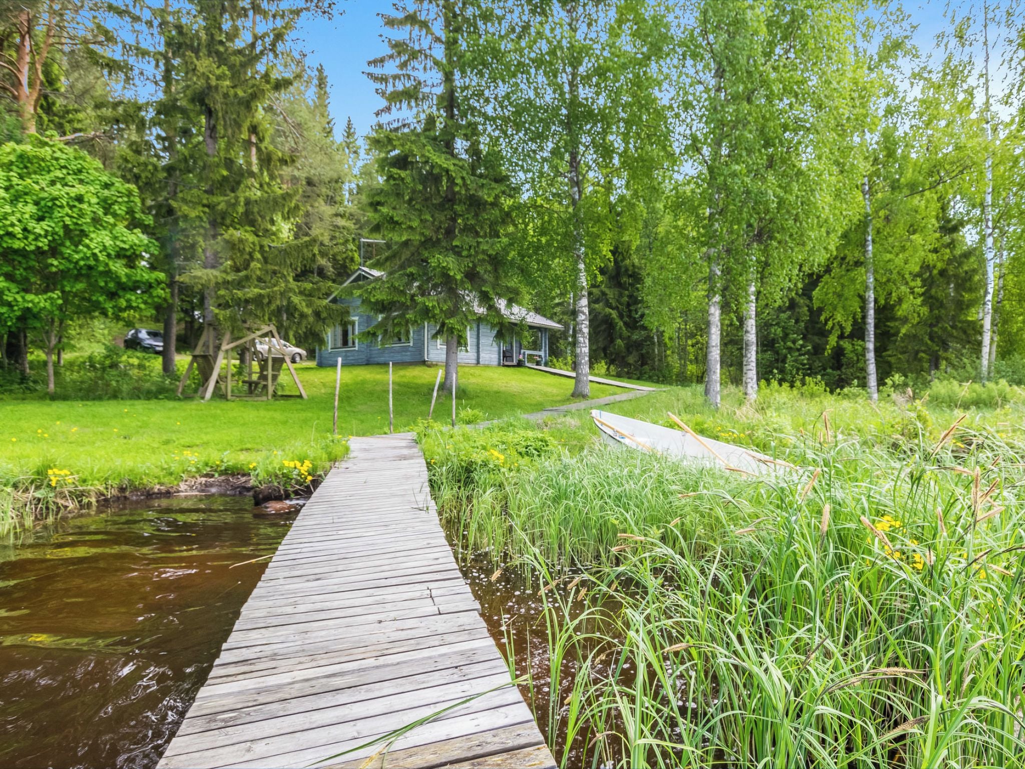 Puinen laituri johtaa talolle, jossa on kuisti. Taloa ympäröi vehreä kasvillisuus ja puut.