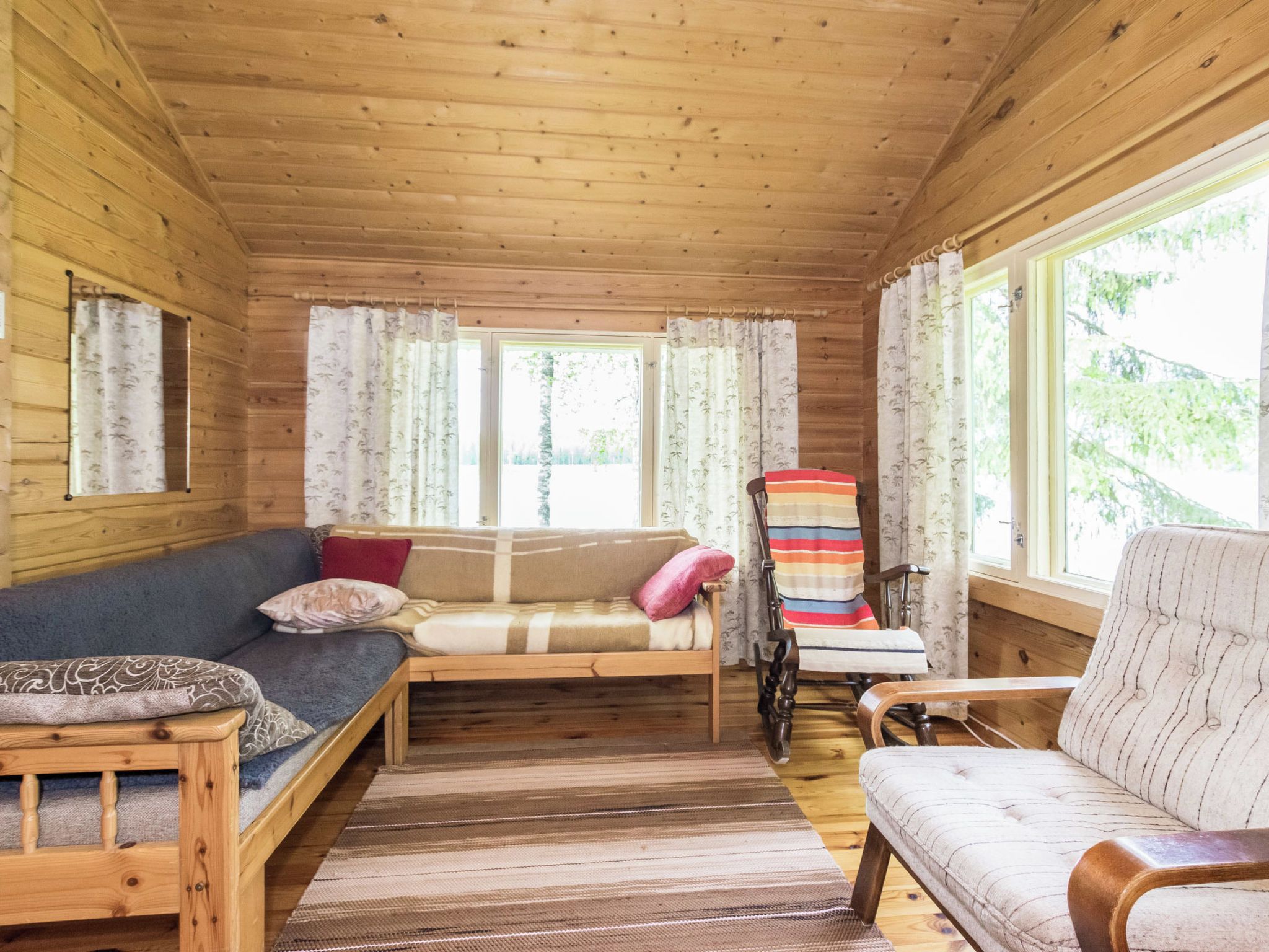 Ein gemütliches Zimmer mit Holzbalkendecke, einer Couch, einem Schaukelstuhl und einem Fenster mit Vorhängen.