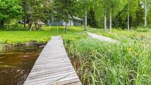 Une jetée en bois menant à une maison entourée d’une végétation luxuriante et traversée par un petit ruisseau.