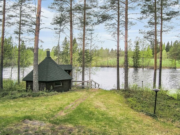 A cabin with a conical roof near a lake surrounded by tall trees.