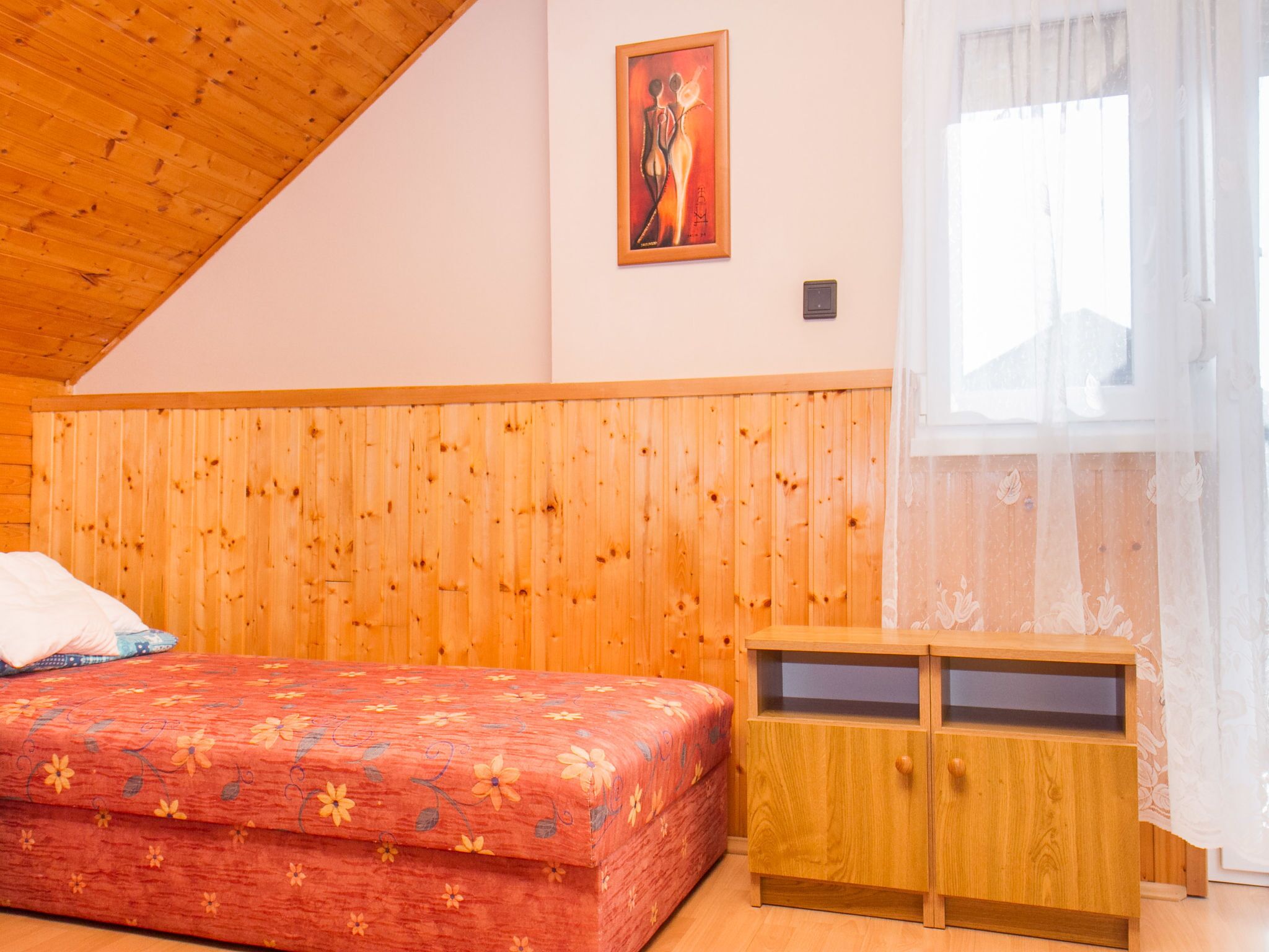 Une chambre avec un plafond en bois, un lit recouvert d’une couvre-lit à motifs floraux, une table de chevet en bois et une fenêtre ornée de rideaux légers.