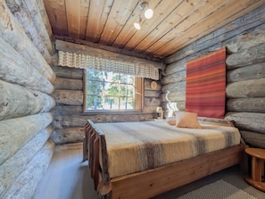 A log cabin bedroom with a wooden bed, a window with a view, and a clock on the wall.