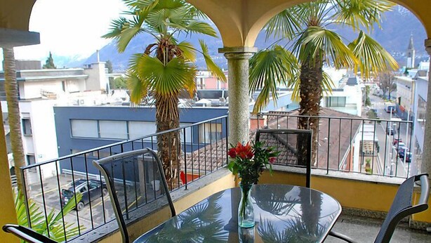 A balcony with a glass table and chairs, surrounded by palm trees and a view of buildings. - Residenza Moro by Interhome (Ascona)